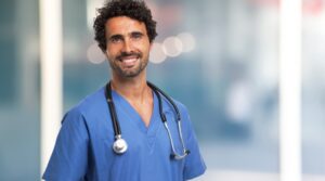 Médico sonriendo, con uniforme azul y estetoscopio alrededor del cuello, en un entorno hospitalario.