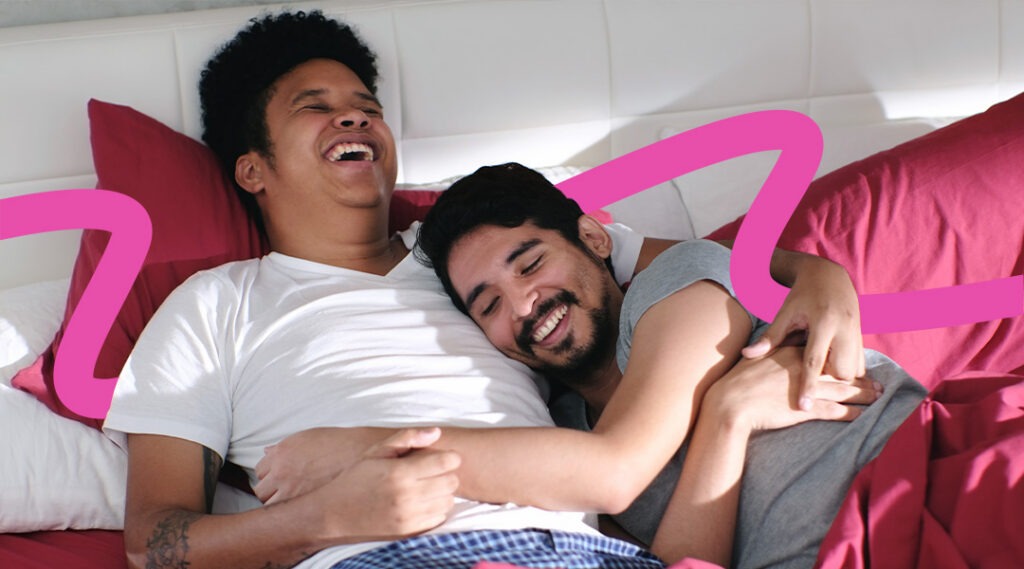 Two men laugh and cuddle while lying on a bed with pink pillows, showing affection.