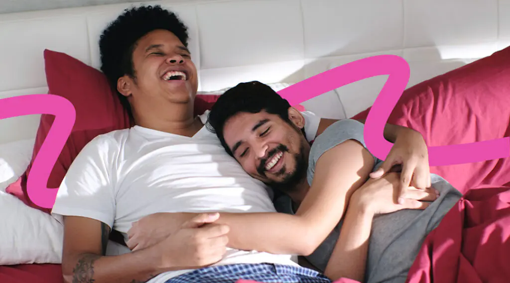Two men laugh and cuddle while lying on a bed with pink pillows, showing affection.