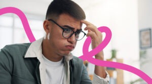 Young man with glasses sits at a desk in an office, looking puzzled and resting his hand on his temple with a pink abstract line in the background.