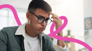Young man with glasses sits at a desk in an office, looking puzzled and resting his hand on his temple with a pink abstract line in the background.